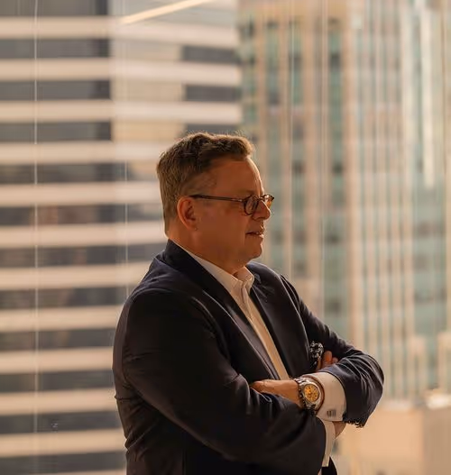 Man with glasses in a suit standing with arms crossed, city buildings visible through the window behind him.