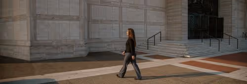 Woman with long hair walking beside a large stone building with steps and metal railings.