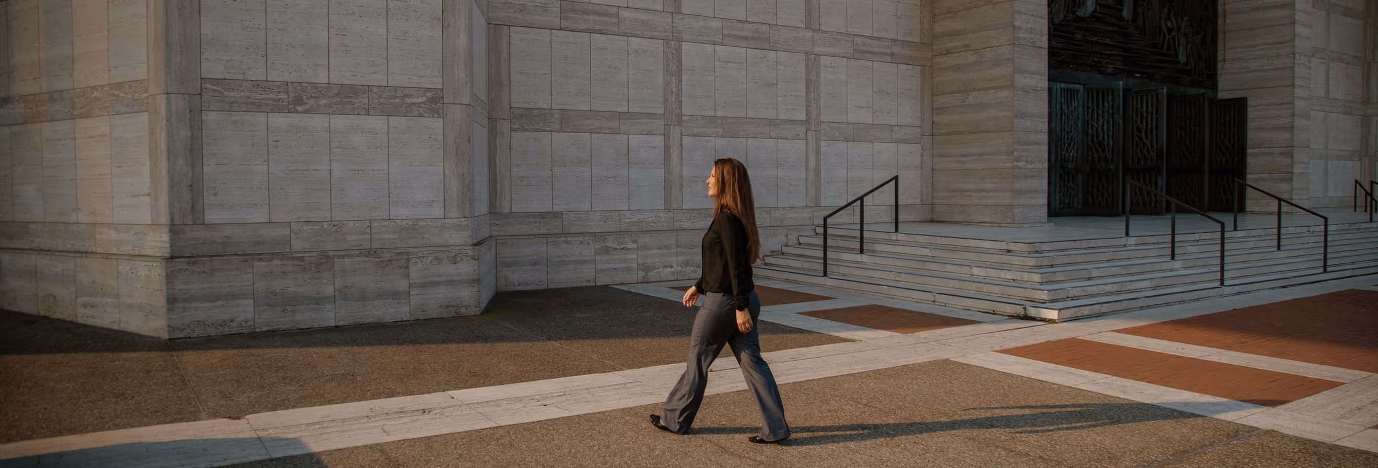 Woman with long hair walking beside a large stone building with steps and metal railings.