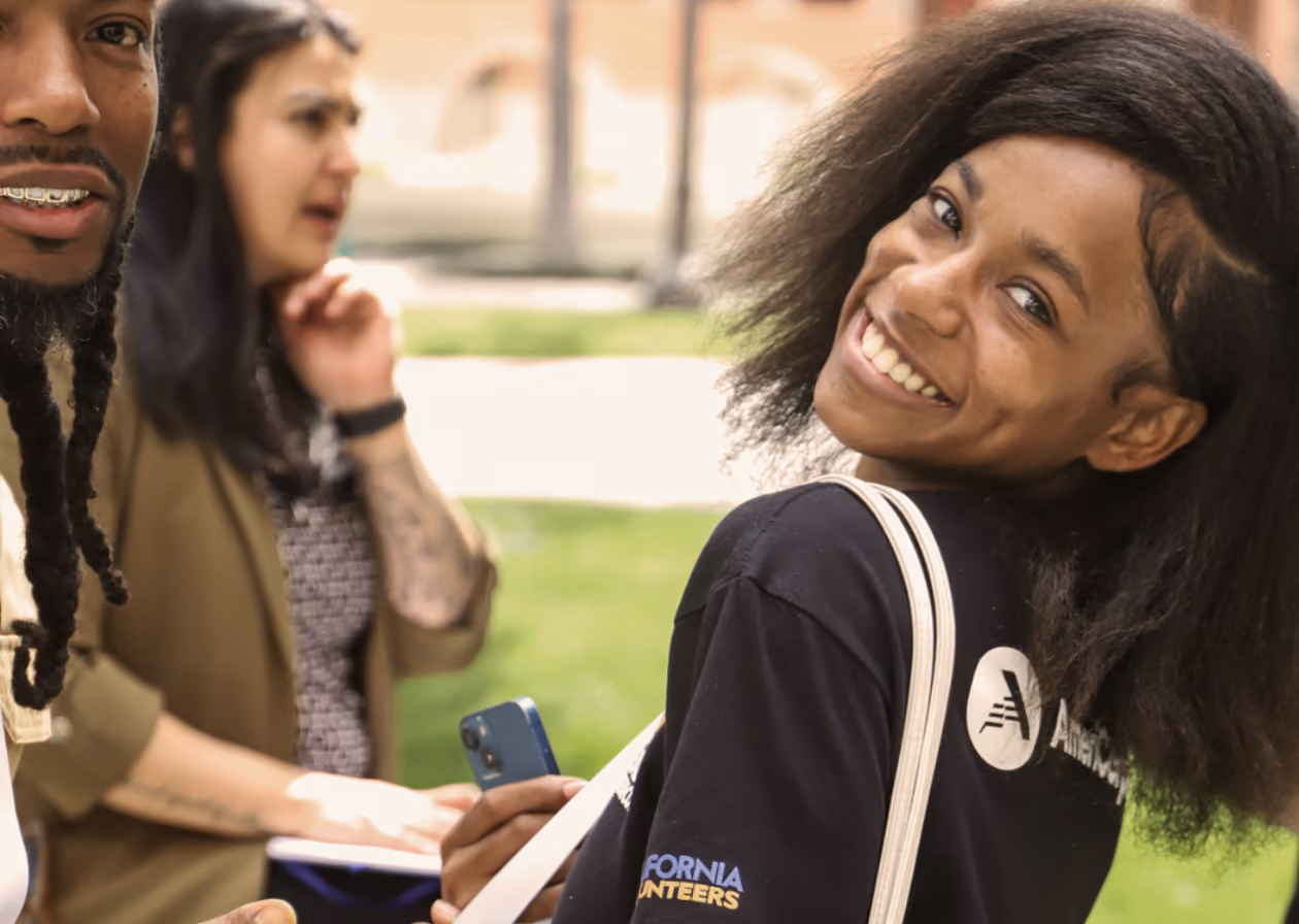 Smiling young woman with natural hair and black shirt looking back, holding phone and tote bag outdoors.