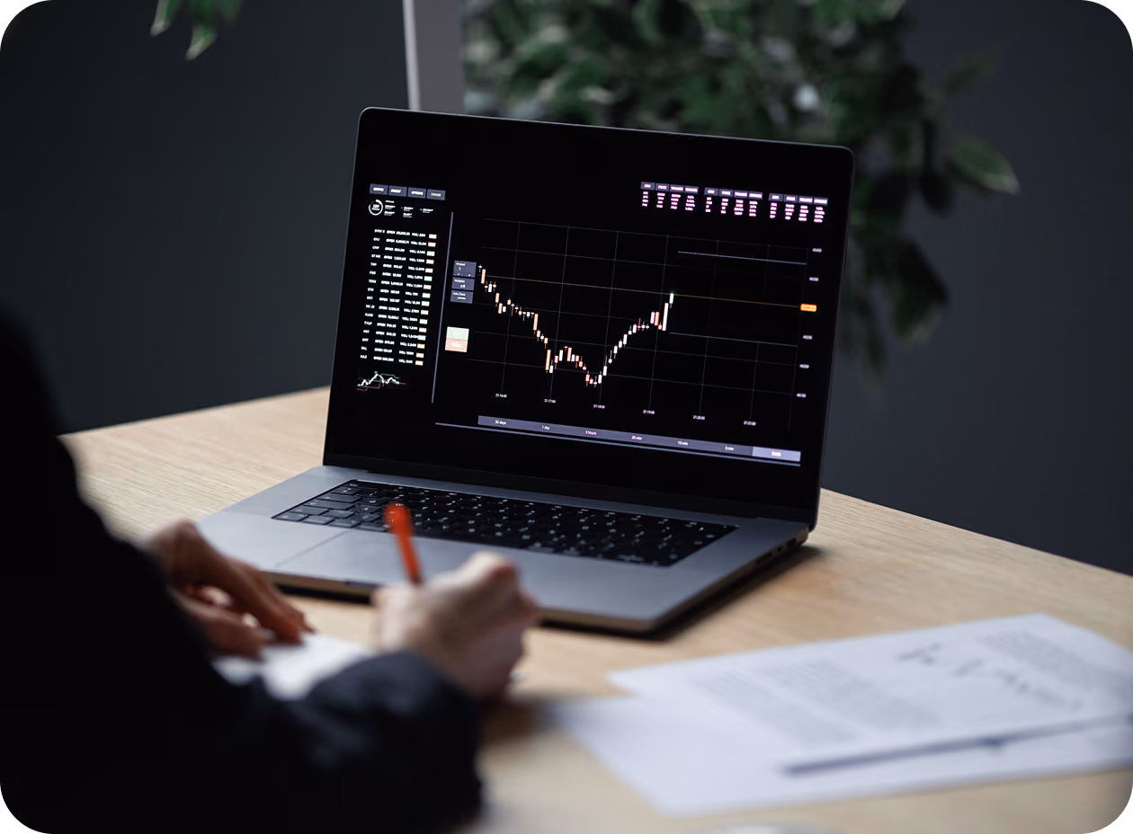 Person writing on paper near a laptop showing a financial chart with data and candlestick patterns.
