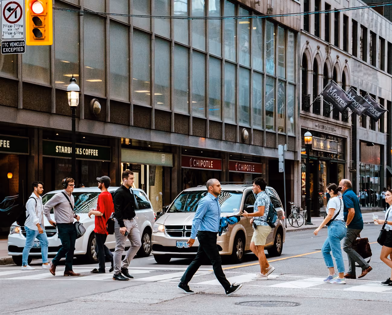 People crossing a busy city street with storefronts.