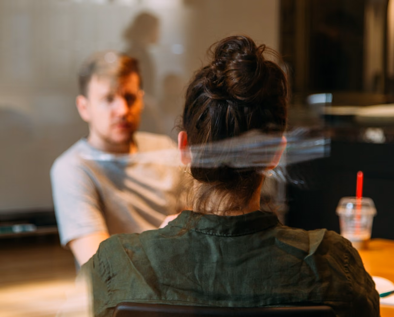 Woman with hair bun facing a blurred man in a casual setting, separated by a clear barrier.