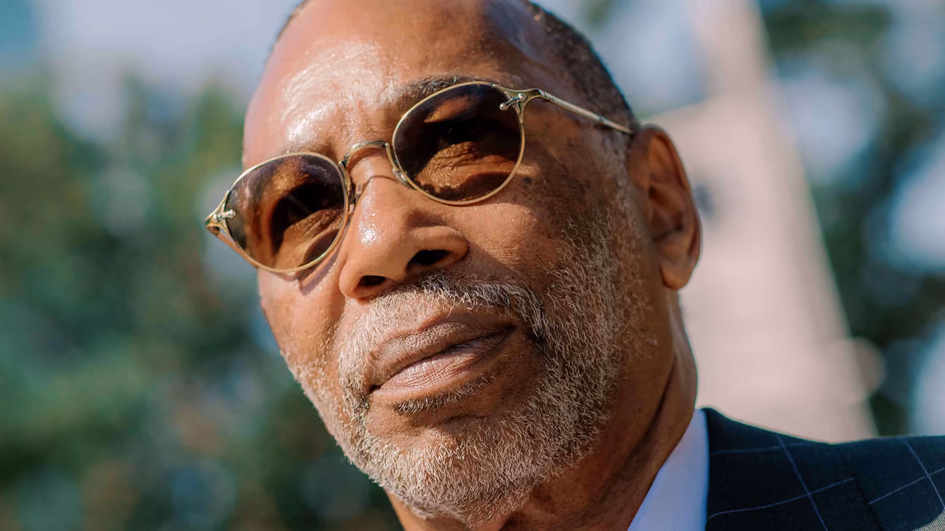 Close-up of a mature man wearing round sunglasses and a suit outdoors with blurred trees behind him.