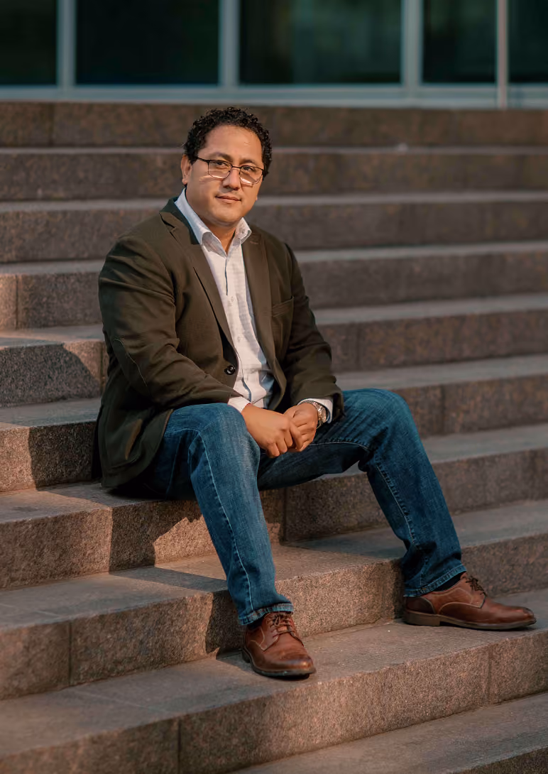 Man wearing glasses, dark blazer, white shirt, jeans, and brown shoes sitting on stone steps outdoors.