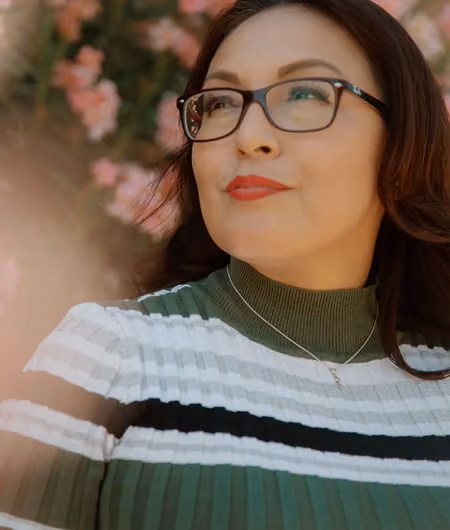 Woman with glasses and red lipstick wearing a striped green and white top, smiling outdoors near flowers.
