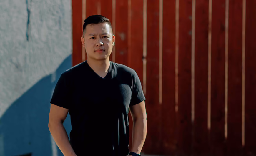 Man wearing black t-shirt standing outdoors with red wooden fence and gray wall background.