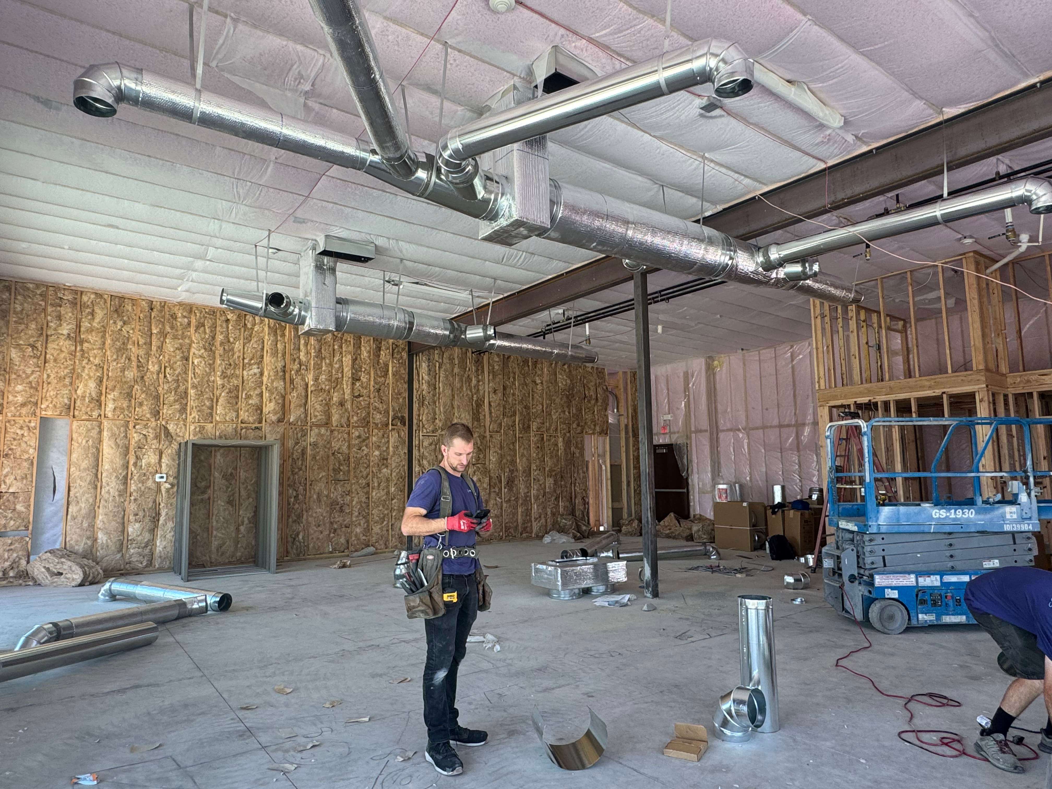 Workers beneath overhead metal duct runs in an unfinished interior.