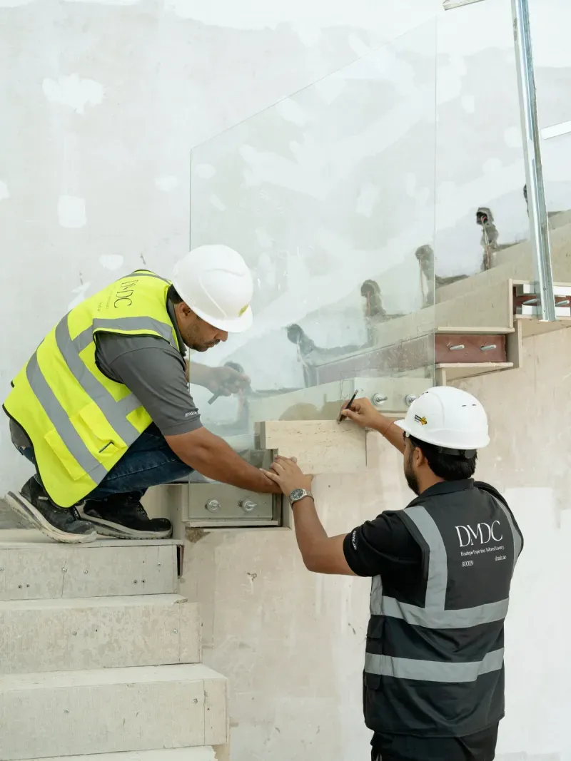 Two DMDC site staff in branded vests and hard hats setting a glass balustrade into a stone stair on site