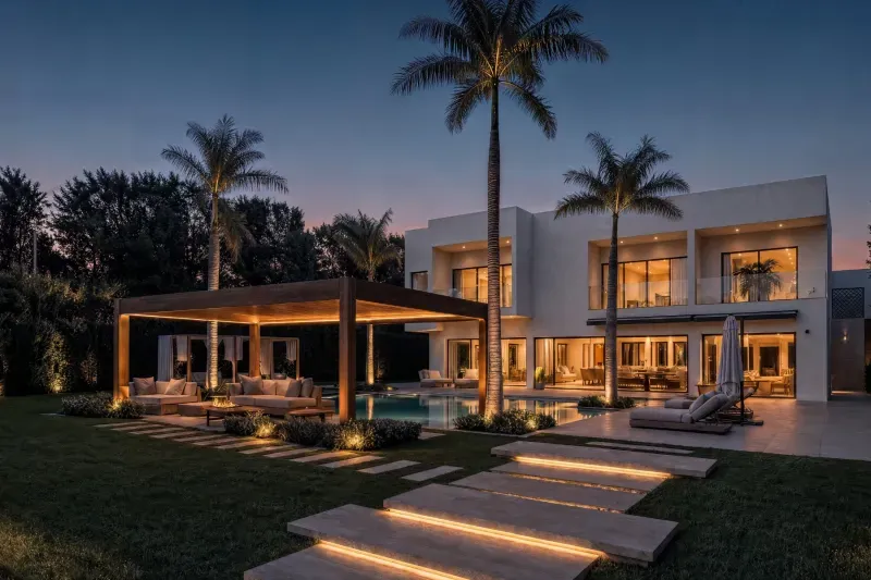 A house in Al Barari, Dubai, at dusk — a lit timber pergola and seating beside the pool, royal palms, and lit stepping stones across the lawn