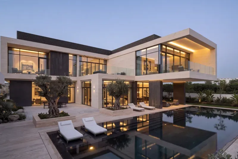 A two-storey travertine house in Mohammed Bin Rashid City at dusk — cantilevered upper floor, olive trees in stone planters and a black reflecting pool