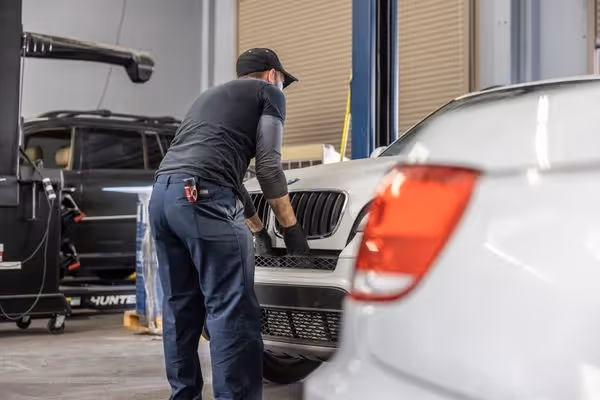Mechanic wearing gloves inspecting the front grille of a silver car in a garage.