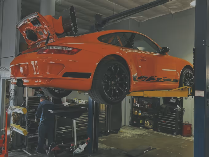 Orange sports car raised on hydraulic lift in a mechanic's garage with its rear engine open.
