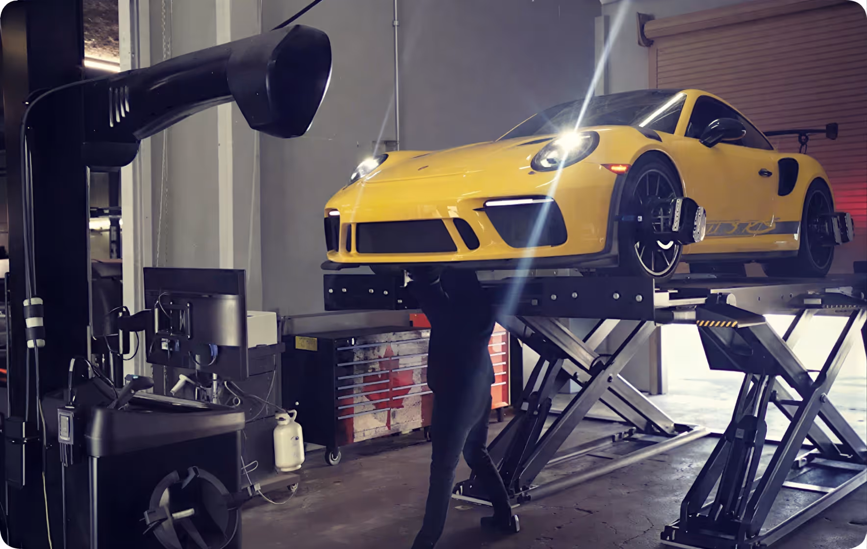 Yellow sports car lifted on platform with mechanic working underneath in a garage.
