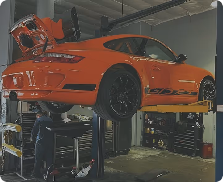 Orange sports car lifted in a garage with rear hatch open and a mechanic working nearby.