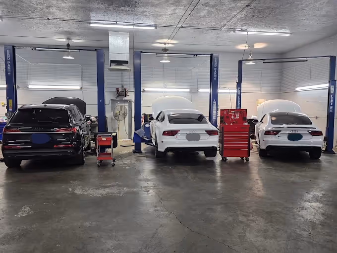 Three Audi cars in a garage service bay with open hoods and tool carts nearby.