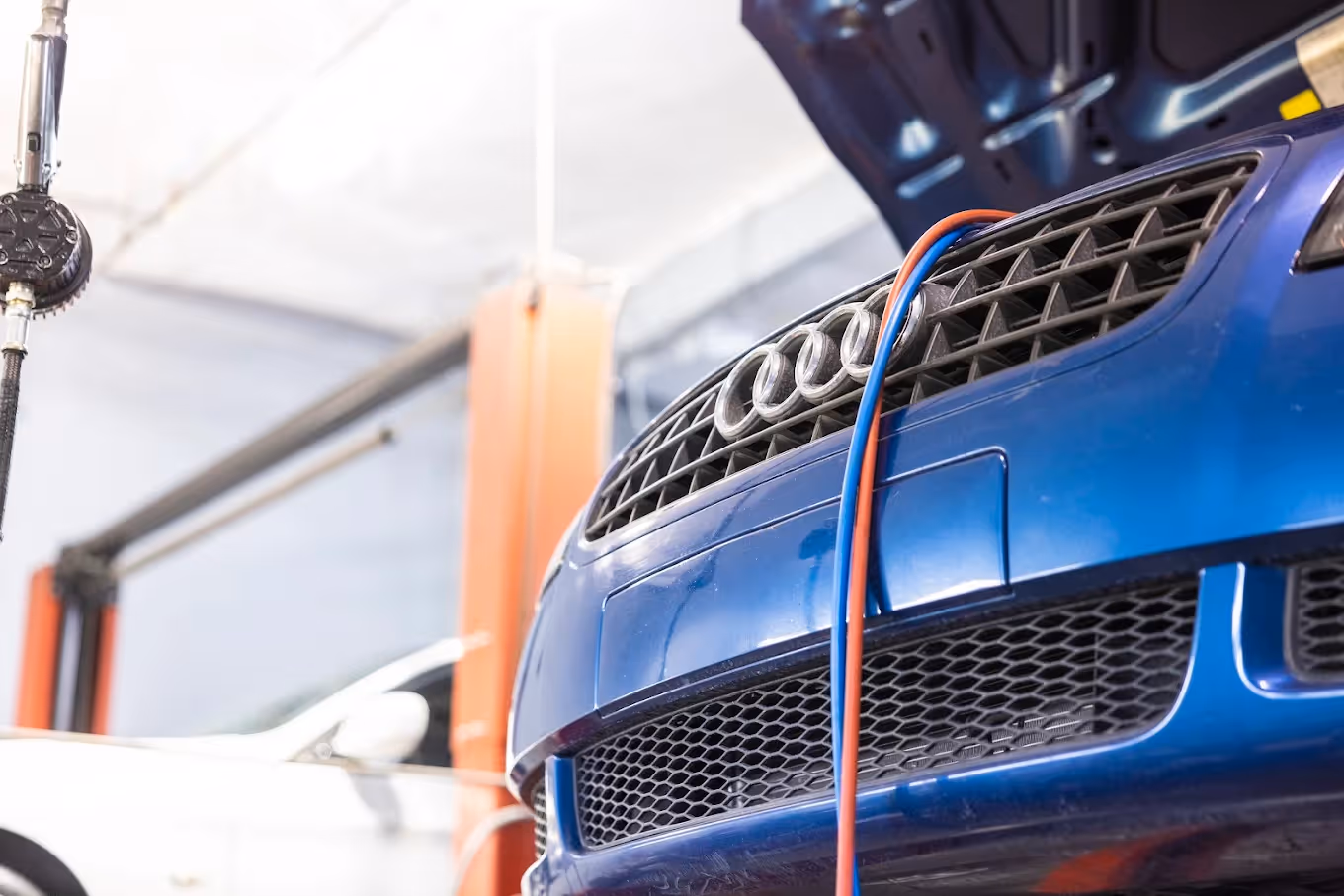 Blue Audi car with hood open and cables attached to the front grille in a garage.