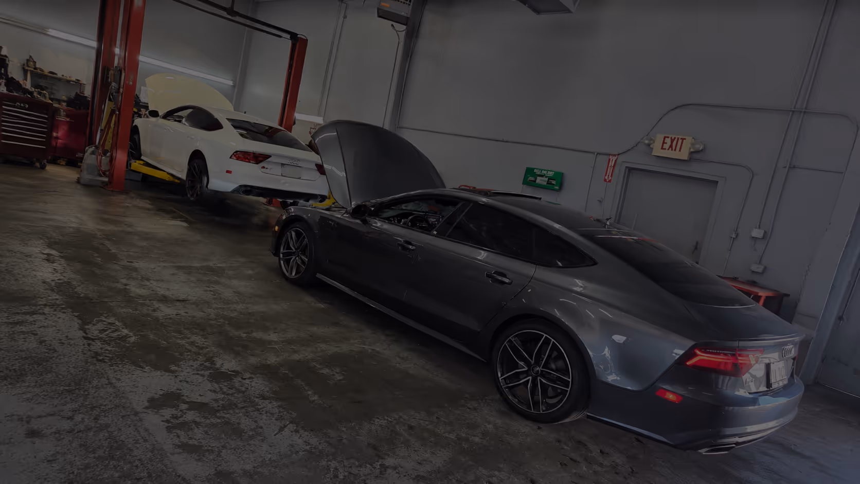 A gray Audi sedan with its hood open inside a garage with a white car on a lift.