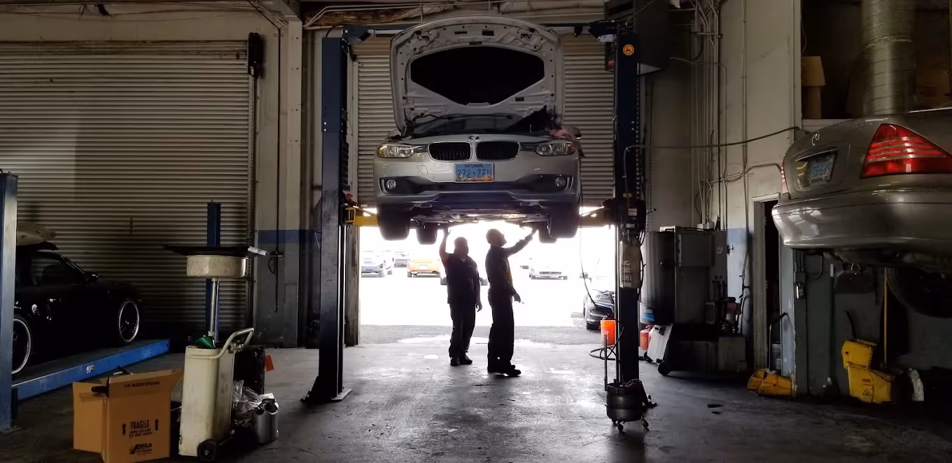 Two mechanics inspect a silver car with its hood open lifted on a hoist inside a garage.