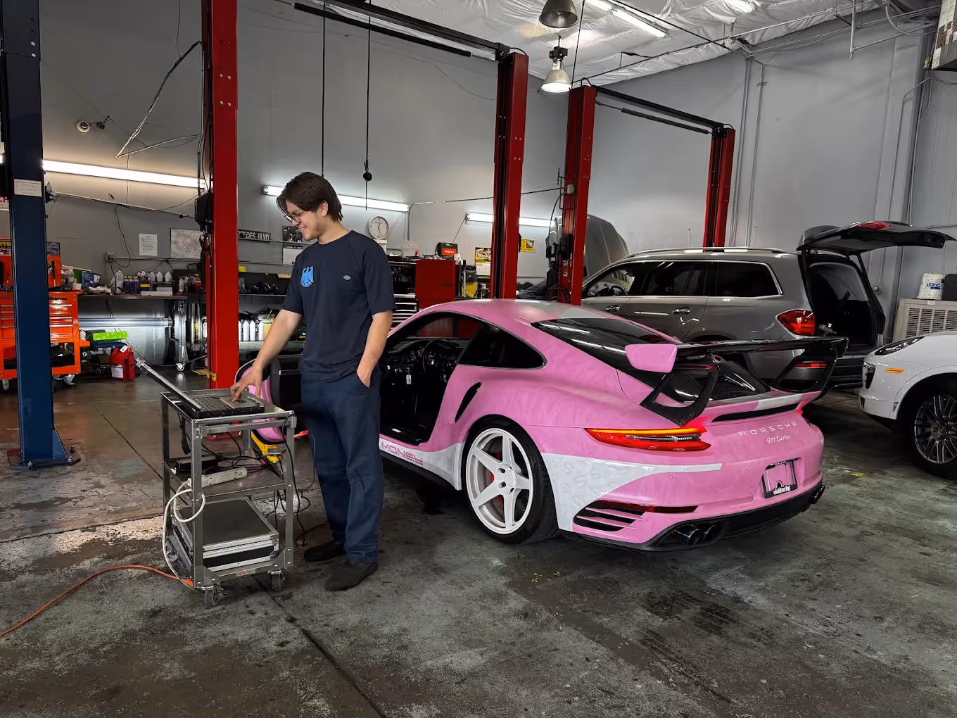 Technician working on laptop next to pink and white Porsche 911 Turbo in a garage.