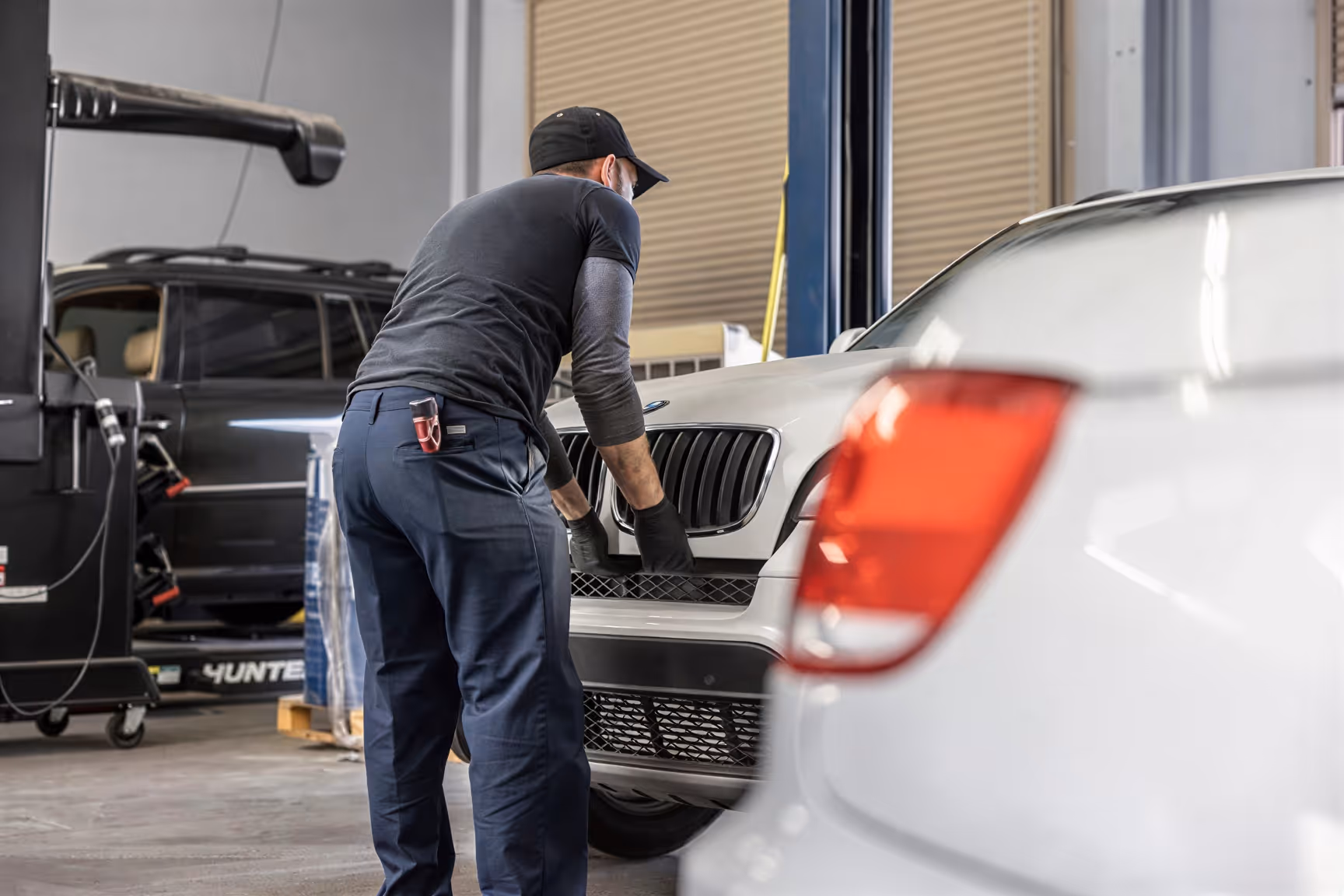 Mechanic in a black cap working on a silver car's front grille in an indoor auto repair shop.