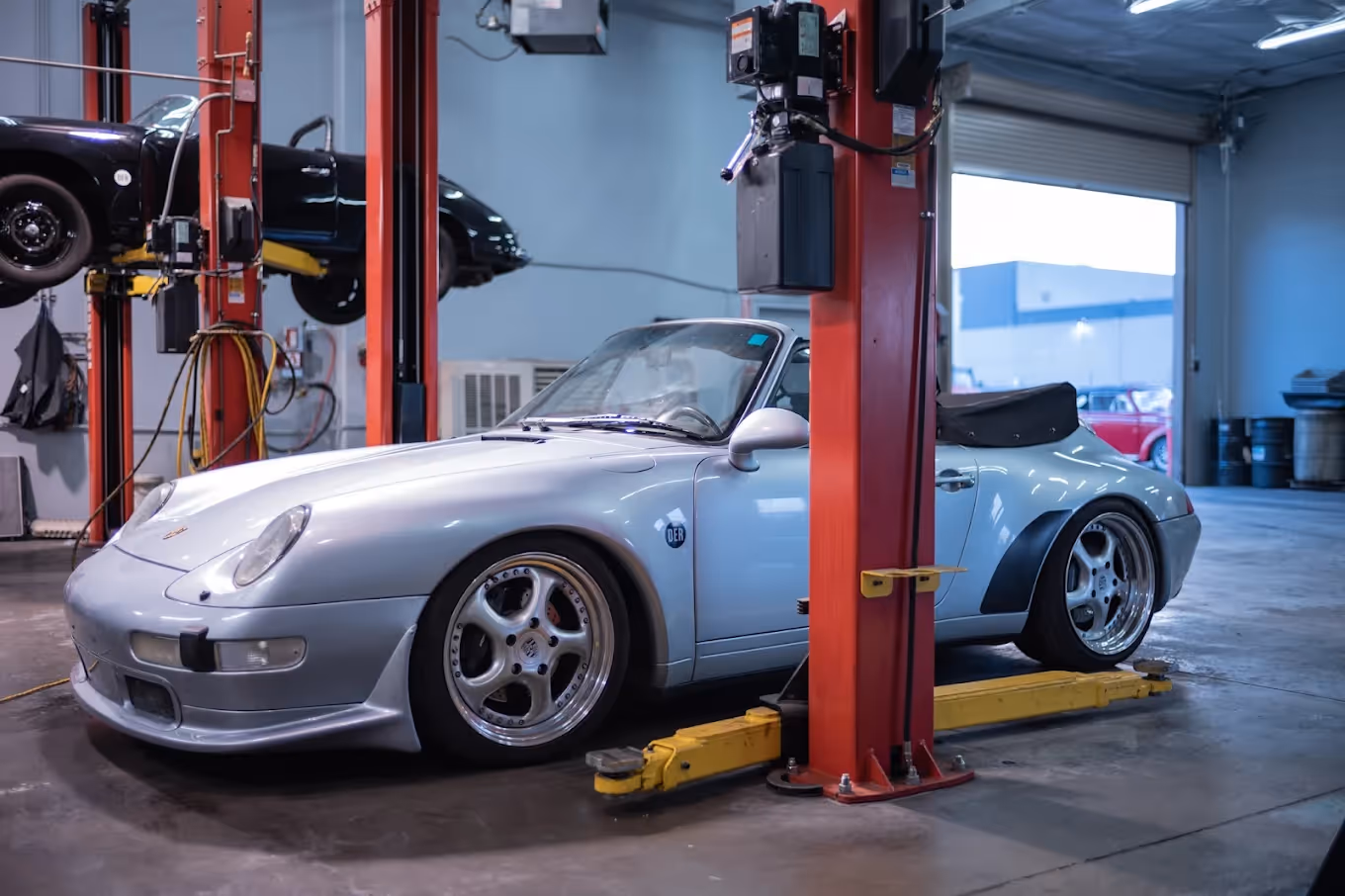 Silver Porsche convertible parked inside a garage with another car on a lift in the background.
