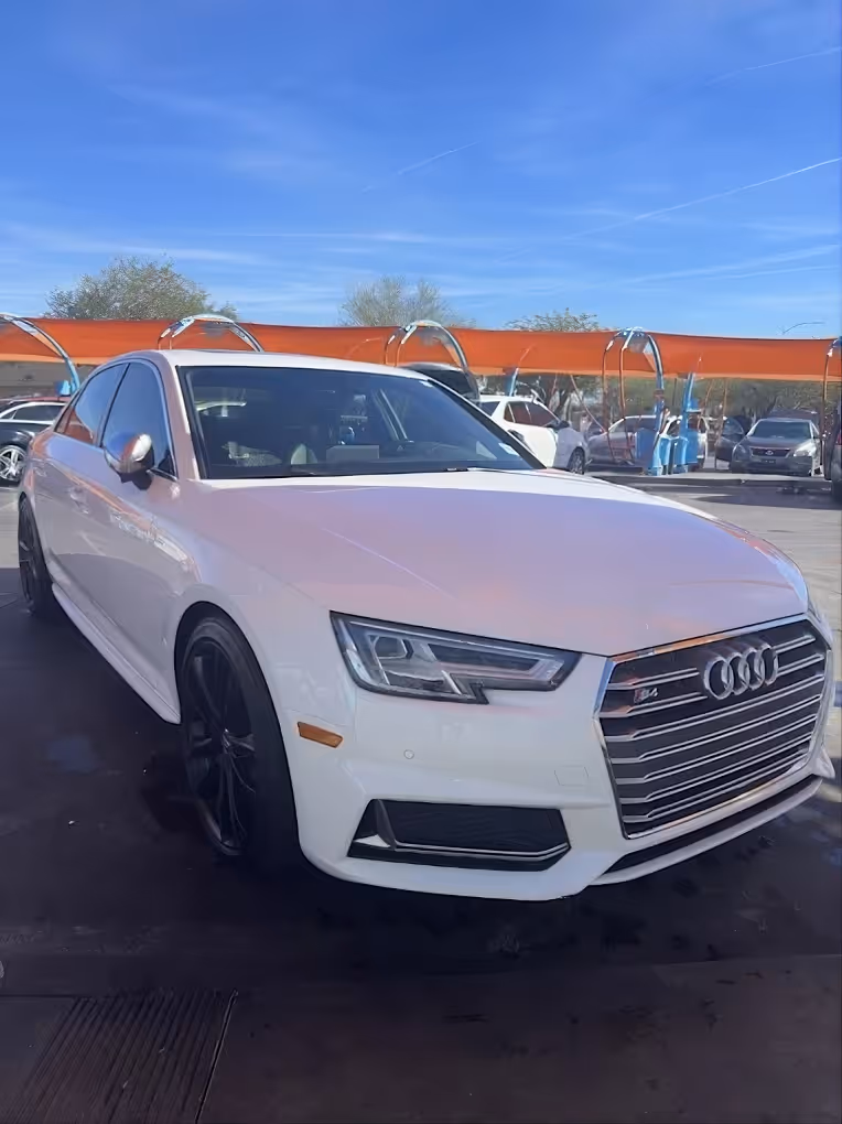White Audi S4 sedan parked outdoors under a blue sky with other cars and orange canopies in the background.