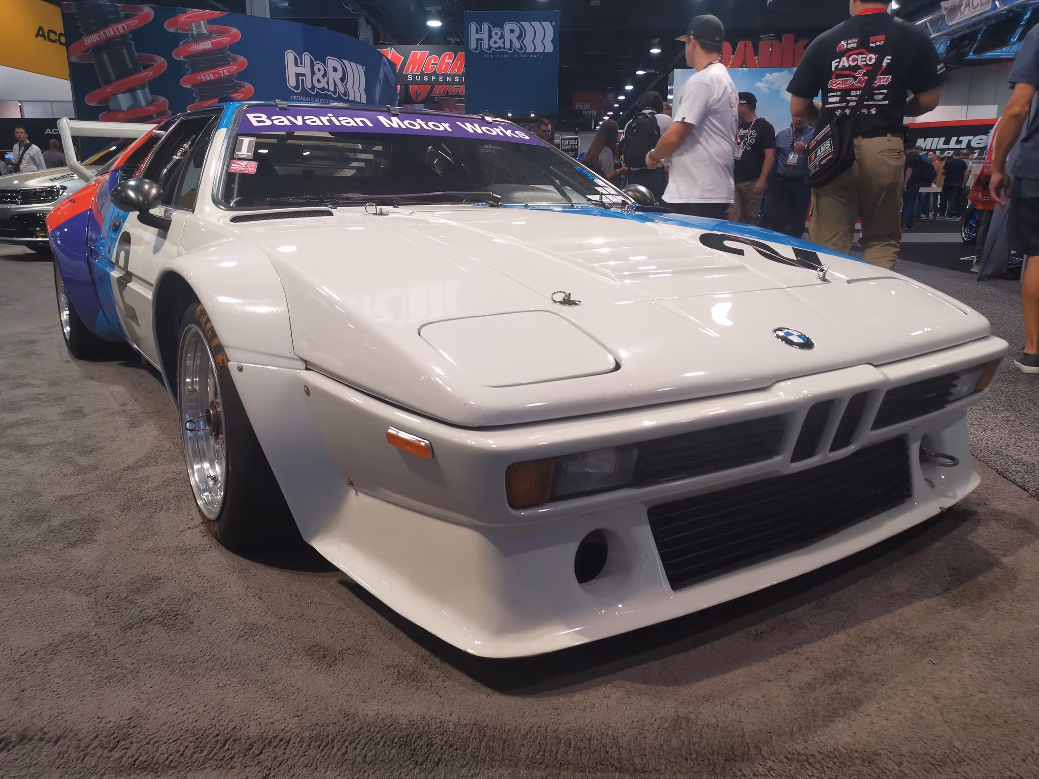 White vintage BMW race car with Bavarian Motor Works banner and colorful side decals at an auto show.