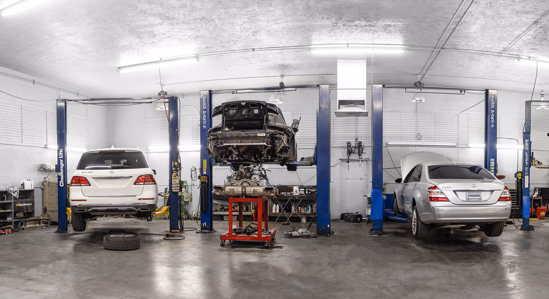 Three cars in a garage on lifts being repaired, one with engine removed on a red cart beneath it.