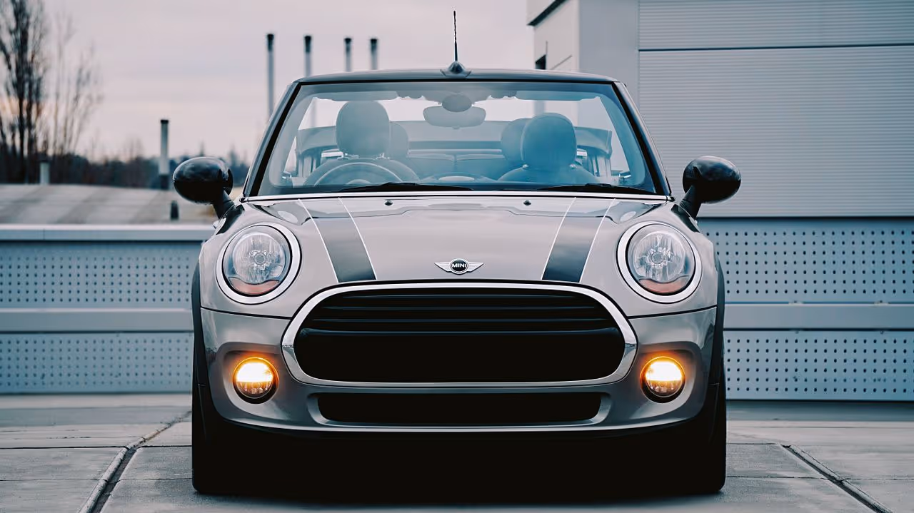 Front view of a silver Mini Cooper with black racing stripes and fog lights on.