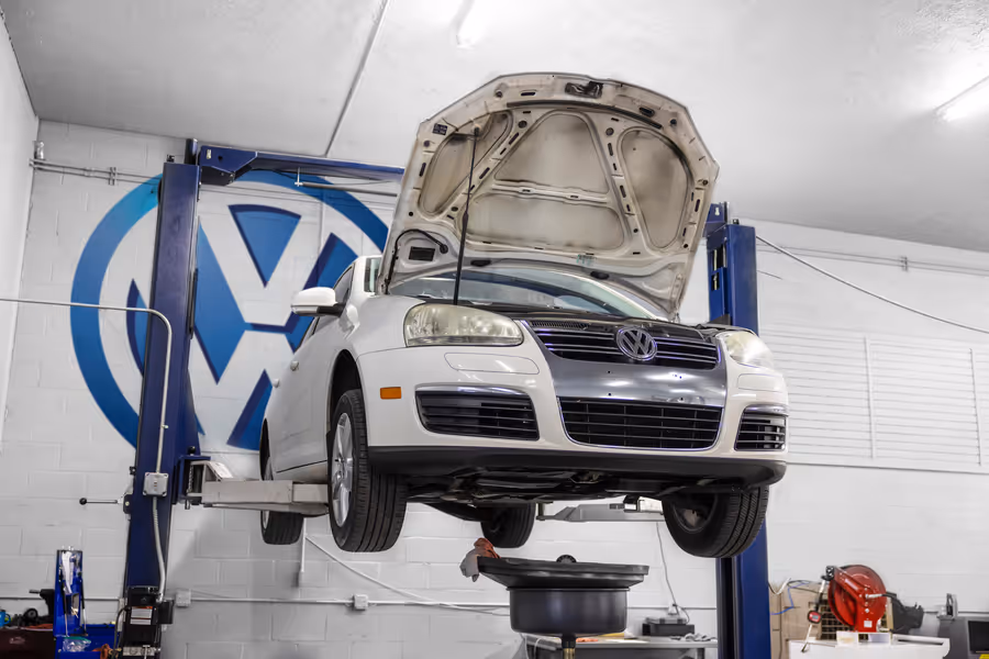 White Volkswagen car with hood open elevated on a hydraulic lift inside a garage with VW logo on the wall.