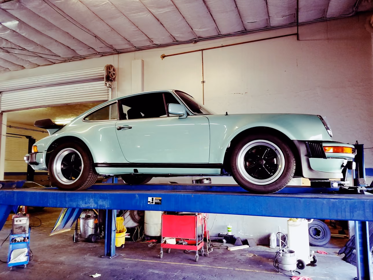 Light green classic sports car on a blue hydraulic lift in an auto repair garage.