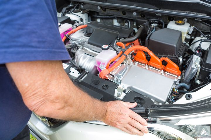 Person inspecting the engine compartment of a hybrid car with visible orange cables and fluid reservoir.