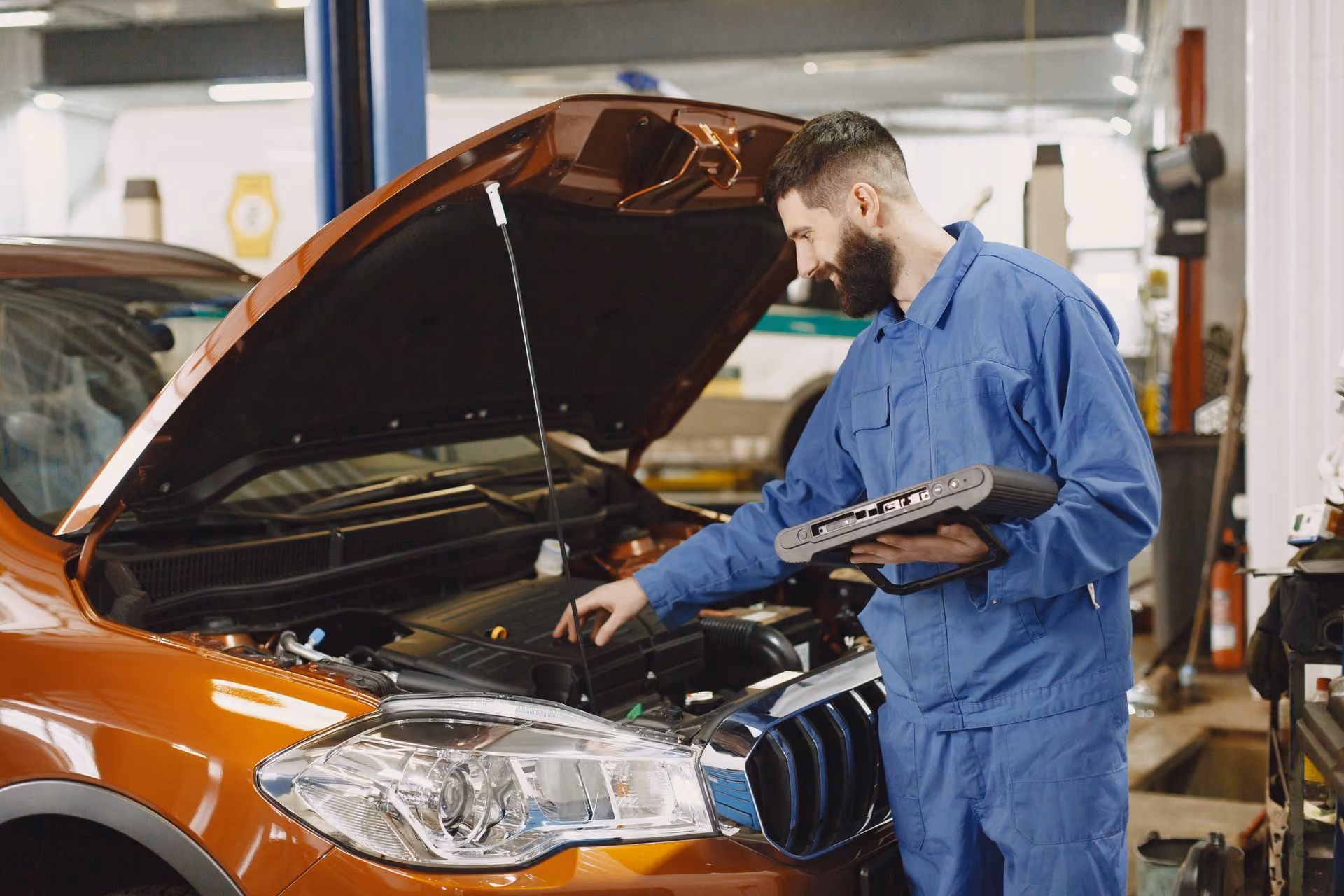 Mechanic in blue coveralls inspecting an orange car engine with a diagnostic device in a garage.