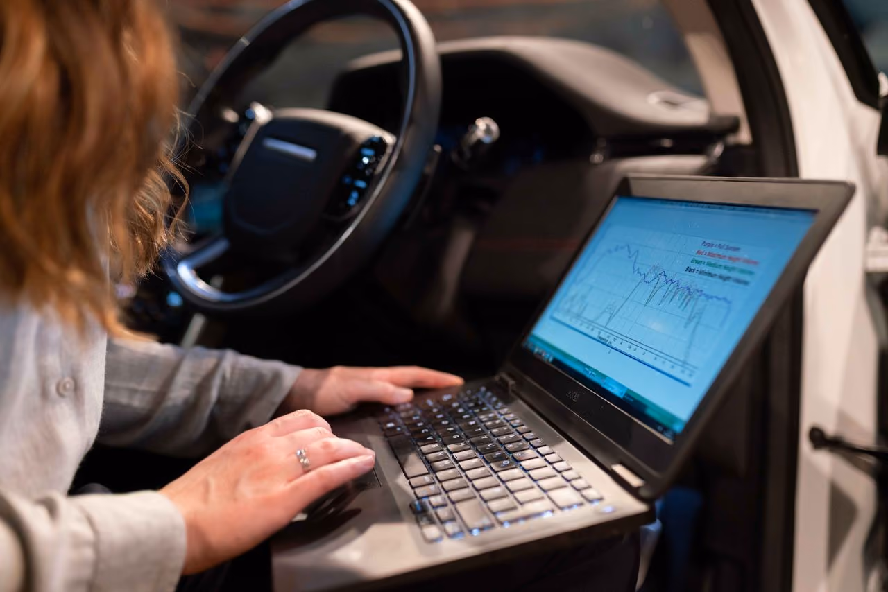 Person using a laptop with diagnostic charts inside a vehicle near the steering wheel.
