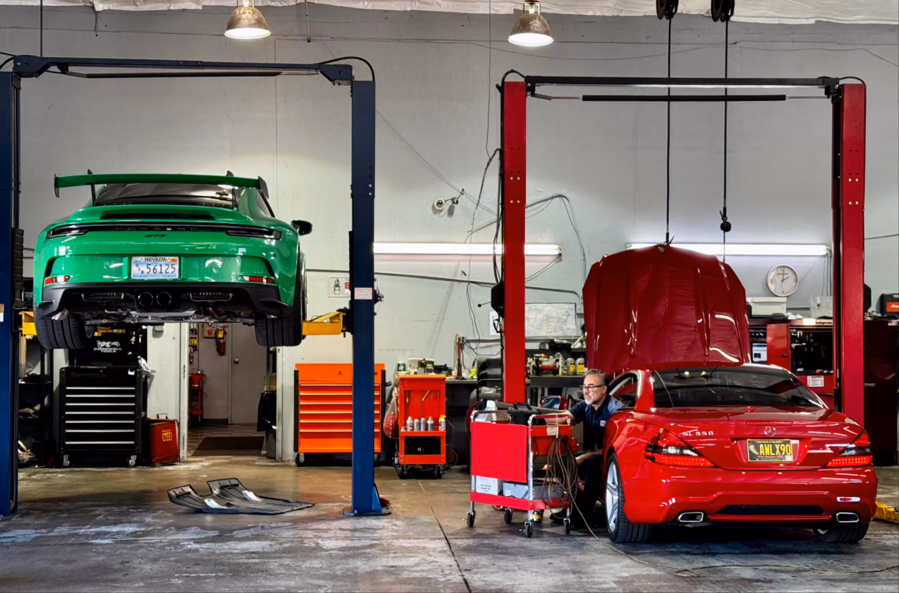 Green car lifted on a hoist and red car with open hood in a mechanic shop with a technician working.