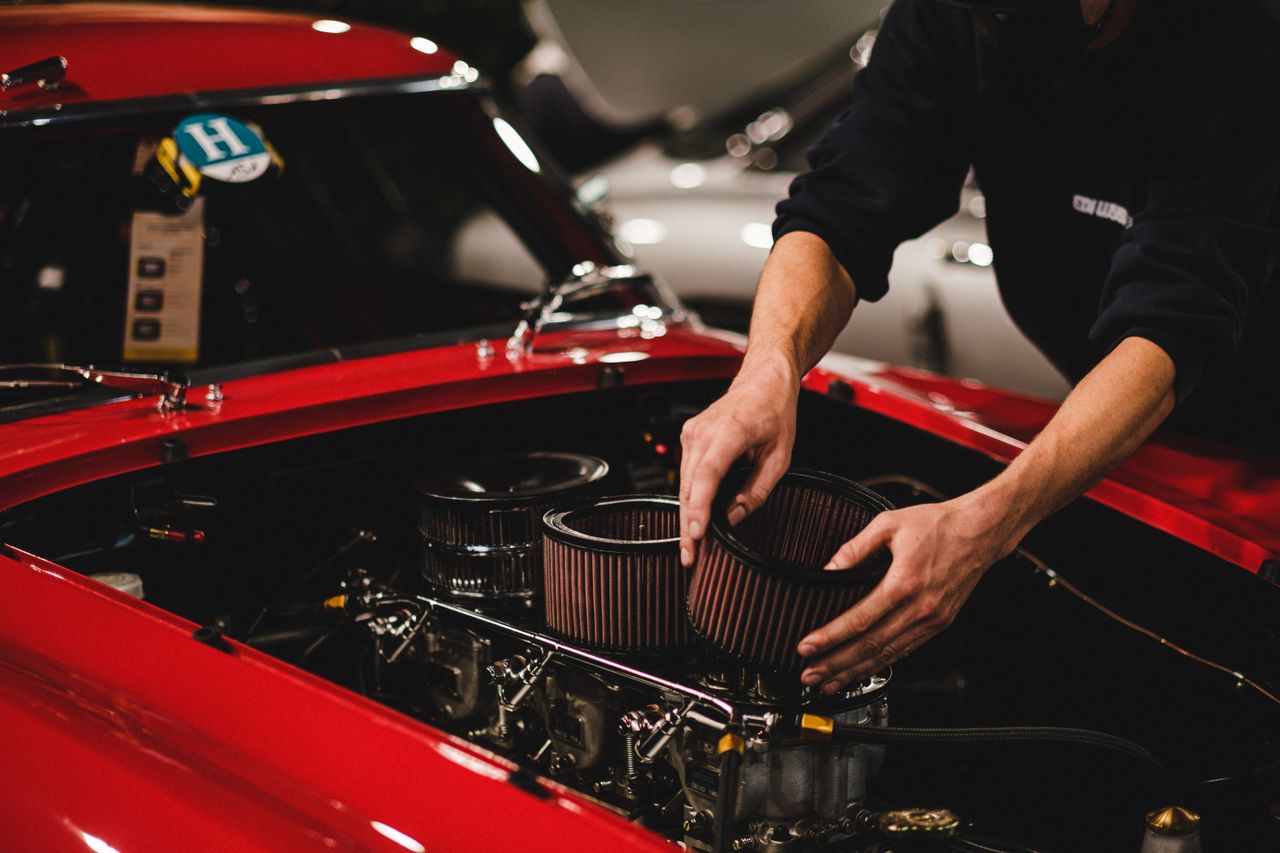 Person's hands working on air filters inside the engine compartment of a red car.