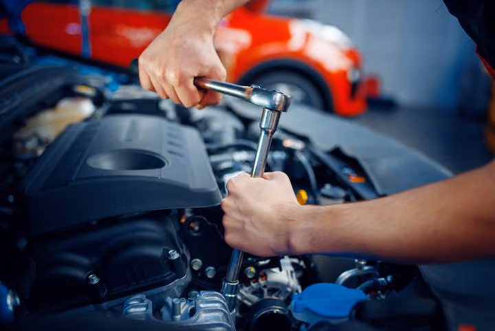 Person using a ratchet wrench to repair a car engine with a red car in the background.