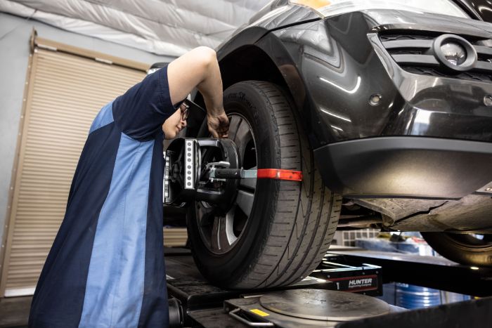 Mechanic in blue uniform adjusting equipment on a raised black SUV's front wheel in a garage.
