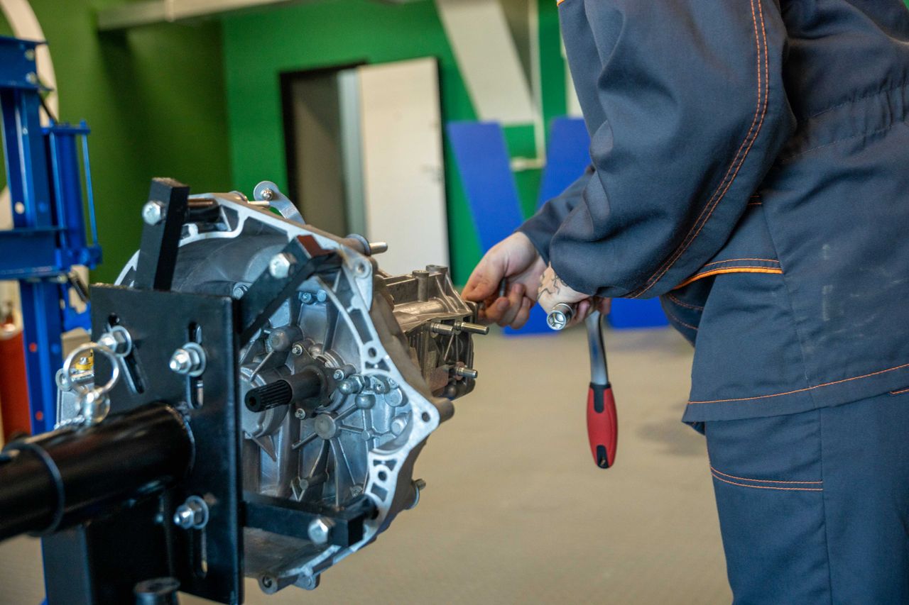 Person in workwear using a wrench on a metal car transmission in a workshop.