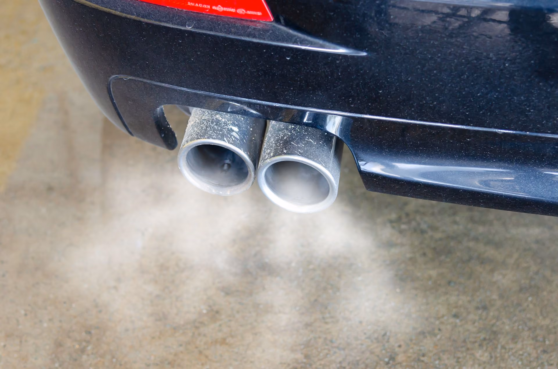 Close-up of car exhaust pipes emitting visible smoke on a concrete surface.