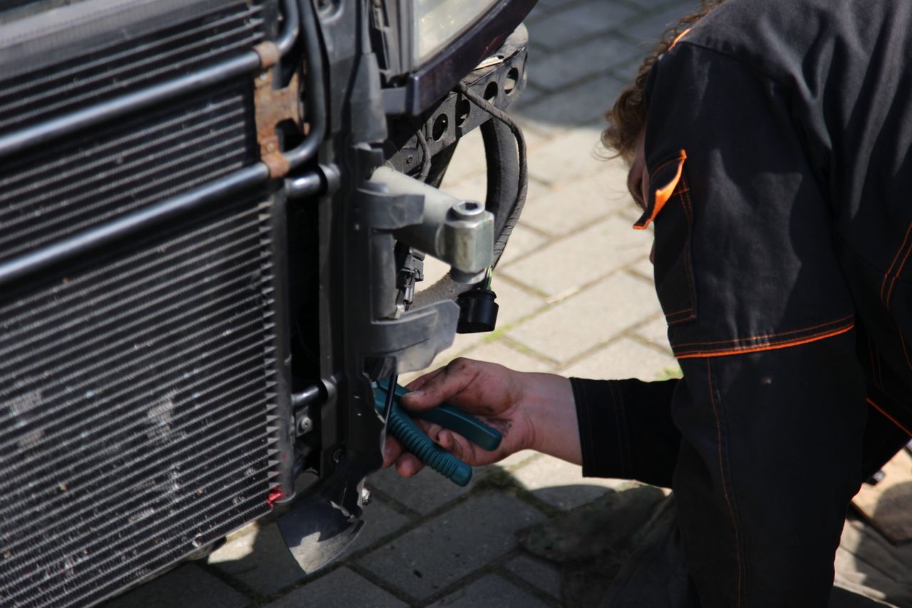 Person using pliers to work on a vehicle's front grill and radiator area outdoors.