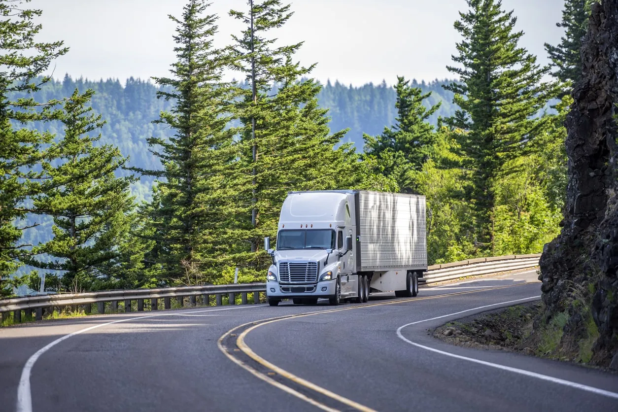 White semi truck driving on a winding road surrounded by green pine trees and forested hills.