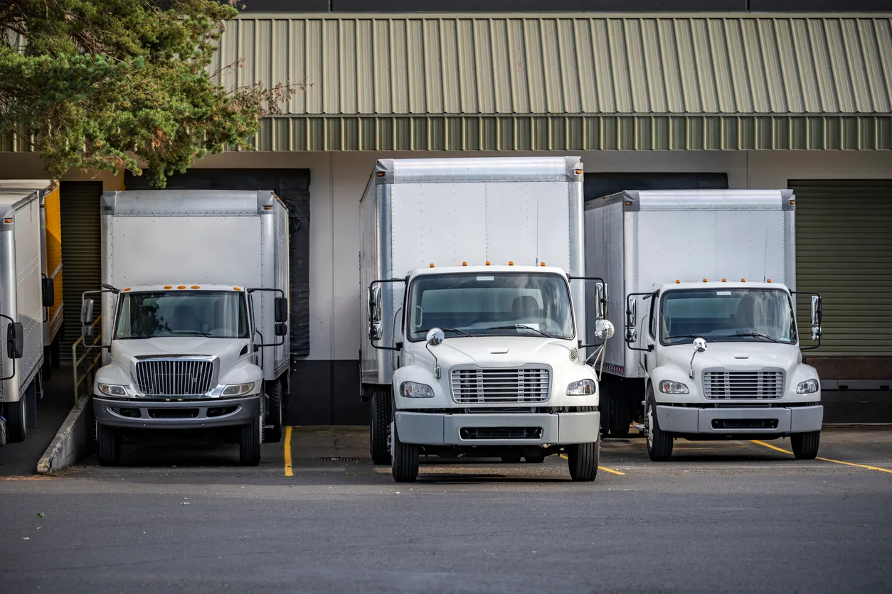 Three white box trucks parked in front of loading dock doors at a warehouse.