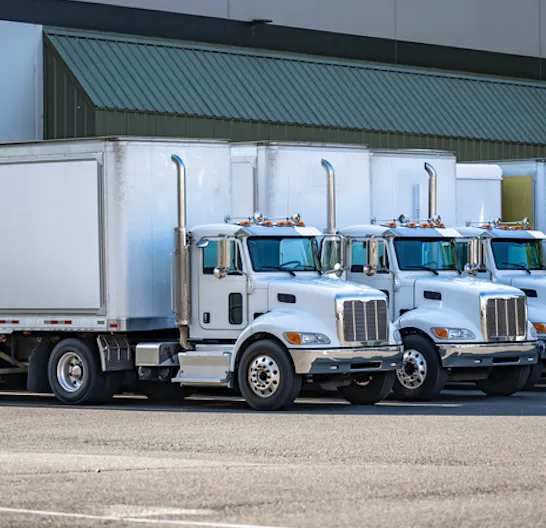 Three white semi-trailer trucks parked side by side in front of a warehouse building.