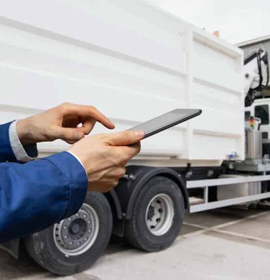 Person in blue jacket using tablet outdoors near a large white truck’s wheel.