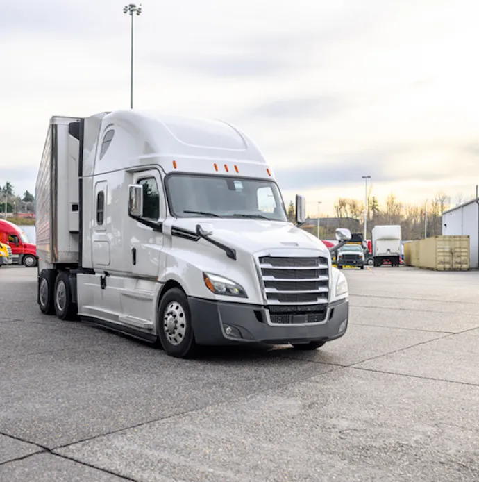 White semi-truck with sleeper cab parked in a lot with other trucks in the background.