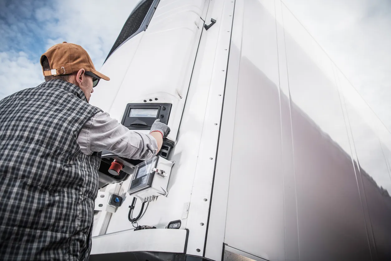Technician in cap and gloves repairing or inspecting a control panel on a white refrigerated truck.