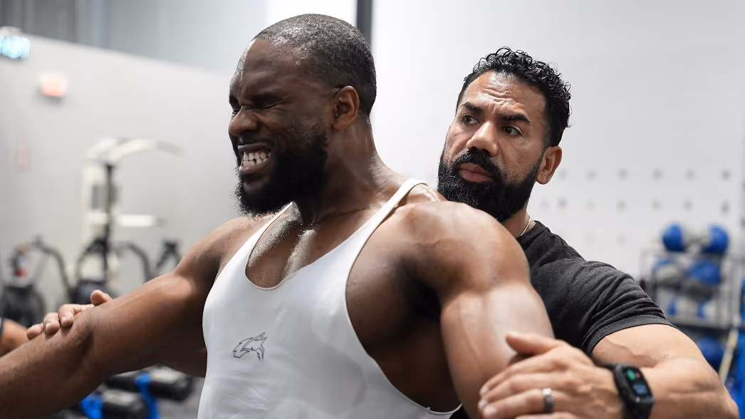 Trainer assists muscular man stretching arms in gym, showing effort and focus during workout.