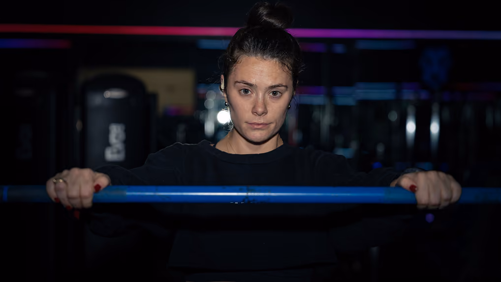 Woman in black shirt gripping a blue bar with focused expression in a dimly lit gym.